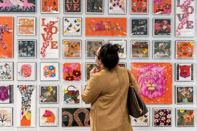 Woman wearing a tan jacket looks at a wall display of framed artworks featuring butterflies, flowers, animals, and brand logos like Gucci and Louis Vuitton.