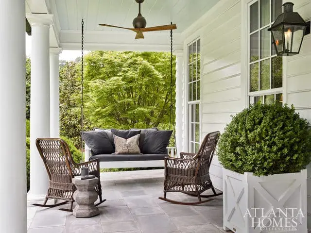 Covered porch with white siding, gray stone tile floor, white porch swing with dark cushions, two wicker rocking chairs, and a large white planter with a green bush. A gas lantern hangs on the wall.
