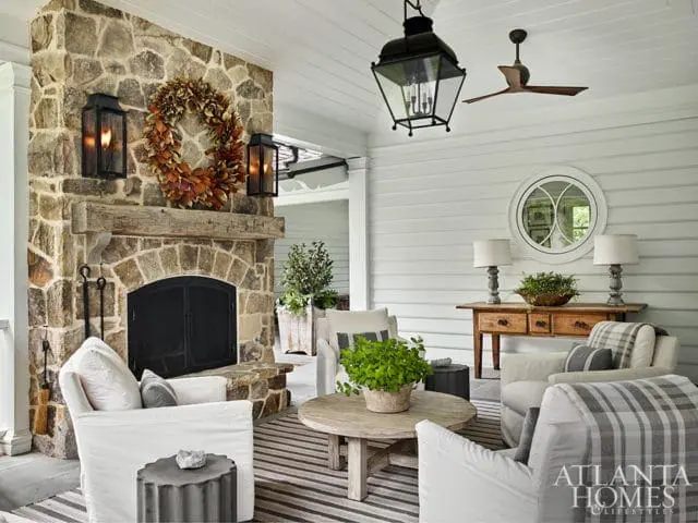 Covered porch with a large stone fireplace, a rustic wooden mantel, and a fall wreath. Seating includes white slipcovered chairs and sofas with plaid throws.