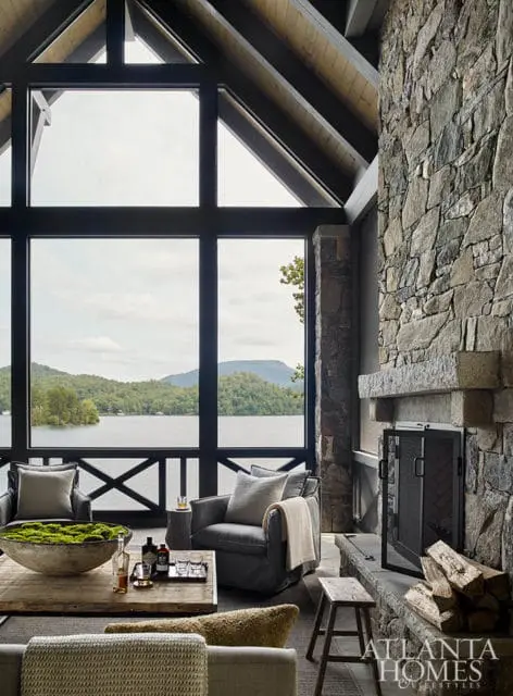 Living room with a large stone fireplace, two gray armchairs, and a rustic wooden coffee table overlooking a lake through a wall of windows.