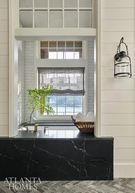 Bathroom with black marble bathtub surround, white subway tile walls, brass fixtures, and a basket of white towels. A window overlooks a body of water.