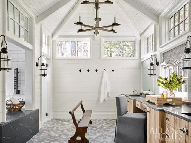 White shiplap walls and vaulted ceiling with wood beams in a room with herringbone tile floors, a wooden bench, and a vanity with wood cabinetry and a stone countertop.