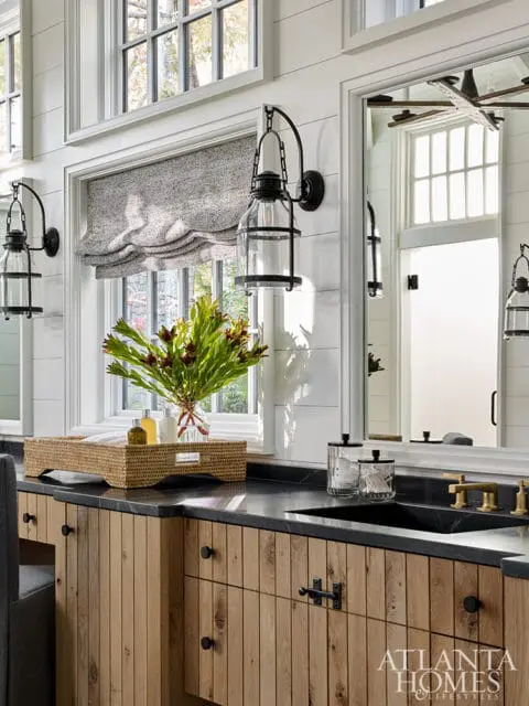 Bathroom vanity with natural wood vertical plank cabinetry, black granite countertop, black sink, and brass faucet. A woven tray with toiletries sits on the counter next to a vase of greenery.