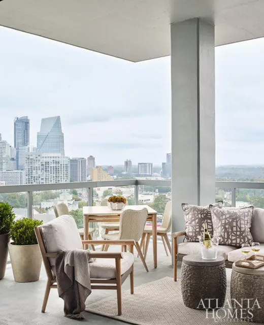 Balcony with light wood furniture including chairs, a sofa, and a dining set, overlooking a city skyline. Pillows with geometric patterns adorn the sofa.