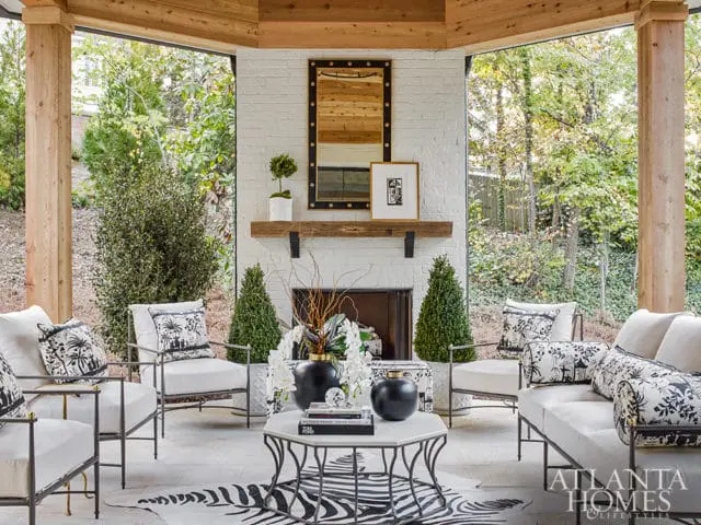 Covered patio with white brick fireplace, wooden beams, white upholstered seating, and black and white patterned pillows. A zebra print rug is on the floor.