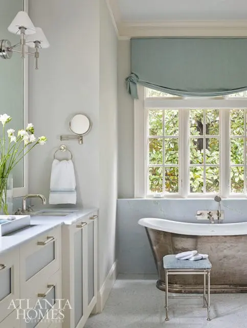 Light blue bathroom with a freestanding copper tub, marble vanity with silver hardware, and a window overlooking greenery.