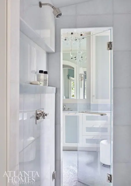 White marble bathroom with glass shower enclosure, vanity with frosted glass cabinets, and a crystal chandelier