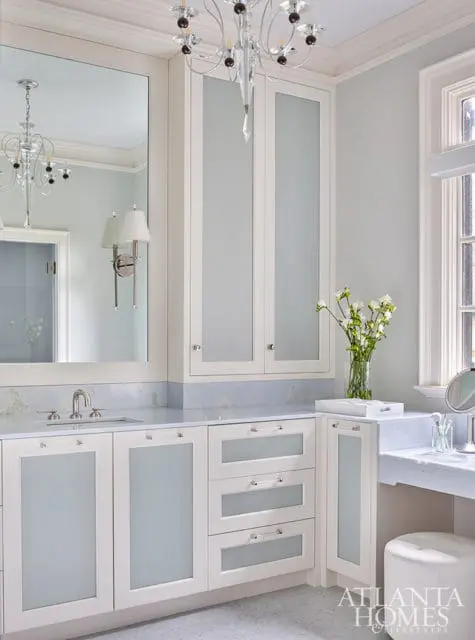 Light blue and white bathroom with white cabinetry, frosted glass doors, marble countertops, and a crystal chandelier.