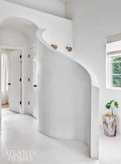 White hallway with curved plaster wall, two doors, and a window overlooking greenery. A log-shaped side table holds a vase with a plant.