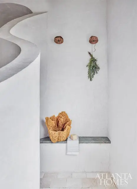 Spa-like bathroom with curved white plaster walls, a stone bench with a large natural sponge and folded towel, and a bundle of dried herbs hanging from the ceiling.