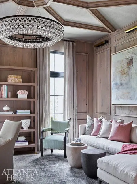 Living room with wood paneled walls, coffered ceiling, crystal chandelier, sofa with pink and gray pillows, and a light green armchair.