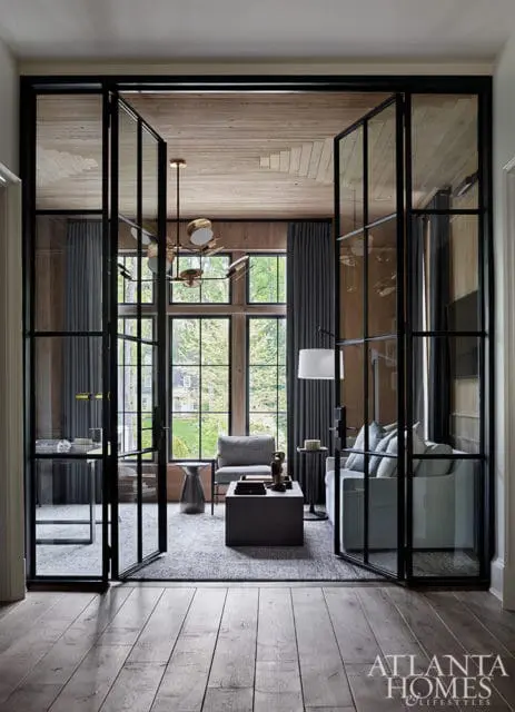 Modern living room with glass-paneled doors, light wood ceiling, gray upholstered furniture, and a large window overlooking greenery.