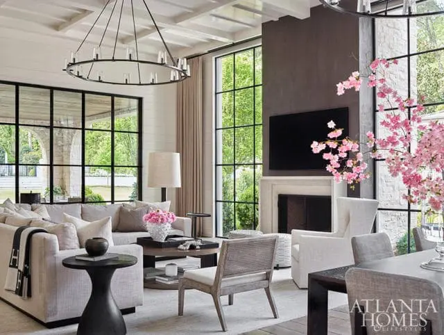 Living room with a large sectional sofa, armchairs, a coffee table, and a fireplace. Large windows overlook a garden. A circular chandelier hangs from the ceiling.