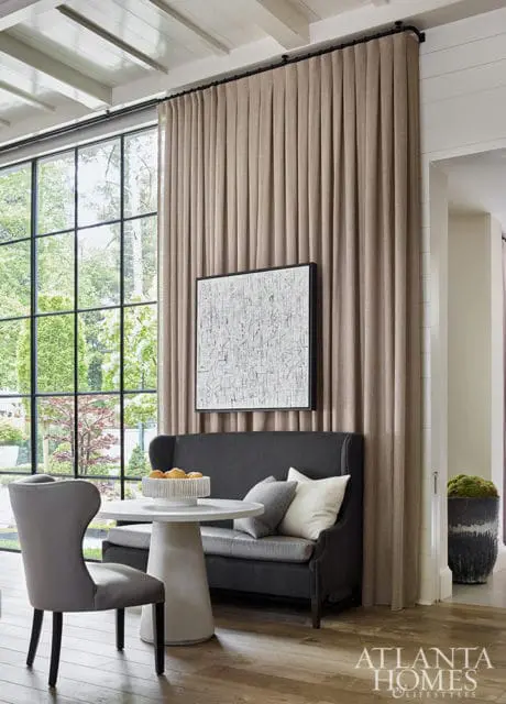 Dining area with a dark gray banquette, white pedestal table with fruit, and gray upholstered chairs. Large window with view of trees and textured artwork on wall.