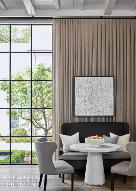Modern dining area with gray upholstered chairs, a dark gray banquette, and a white pedestal table with a bowl of fruit. A large abstract artwork hangs on the wall above the banquette.