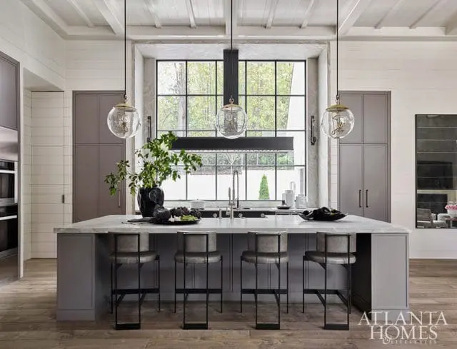 Gray kitchen island with marble countertop, four gray leather bar stools, and a large window overlooking trees. Tall gray cabinets line the walls.