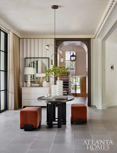 Foyer with a round dark wood table, two rust-colored ottomans, a white dresser, and a sculptural pendant light. An arched doorway reveals a staircase.