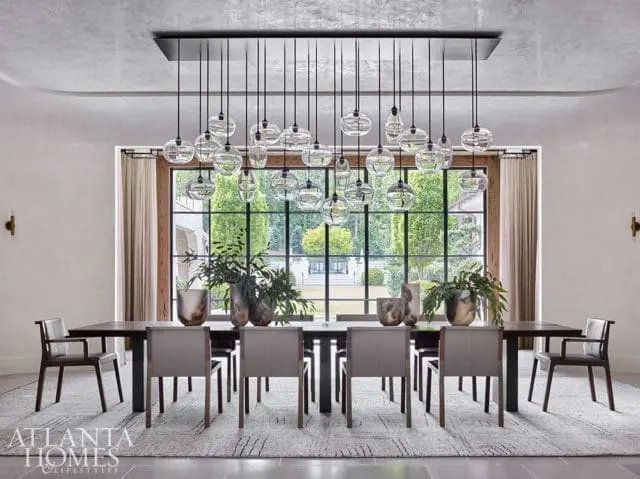 Modern dining room with a long dark wood table, upholstered chairs, and a large window overlooking a garden. A multi-bulb glass pendant light hangs above the table.