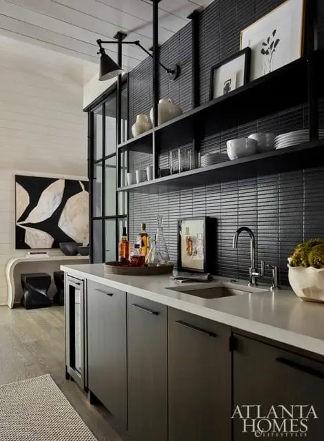 Dark wood bar with white countertop, stainless steel sink, and black tiled backsplash. Open black shelves display glassware, bowls, and decorative items.