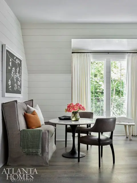 Dining area with shiplap walls, a gray upholstered banquette, marble-top tulip table, and leather chairs.