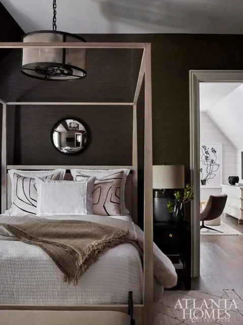 Bedroom with a four-poster bed, dark textured wallpaper, and a view into an adjacent room with a chair and dresser.