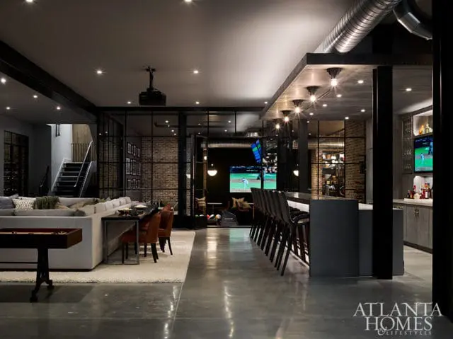 Modern basement entertainment area with a long sectional sofa, bar seating, exposed brick walls, and a large screen displaying a baseball game.