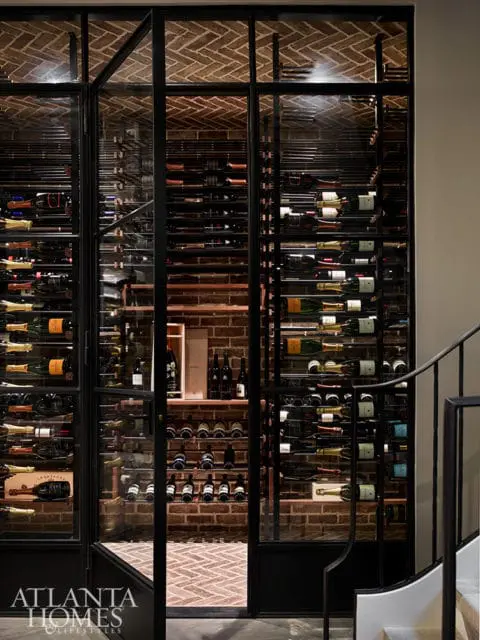 Glass-fronted wine cellar with brick walls and herringbone brick floor, filled with wine bottles on metal racks.