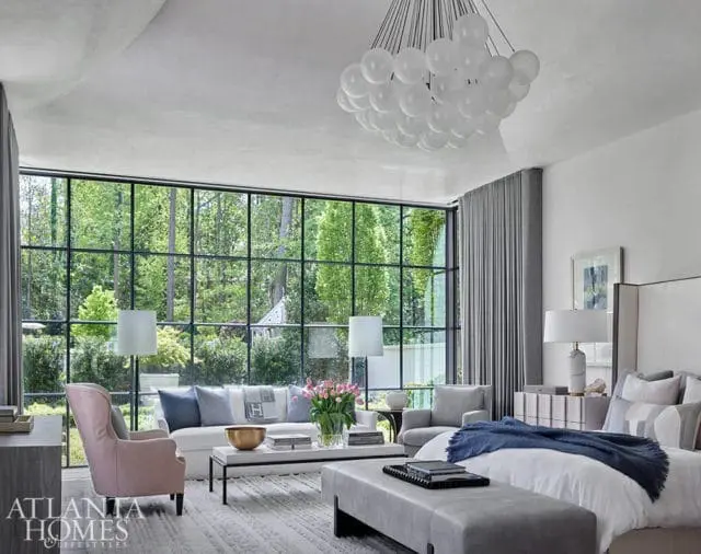 Bedroom with large grid window overlooking trees, white sofa, pink armchair, king bed with blue throw, and modern bubble chandelier.