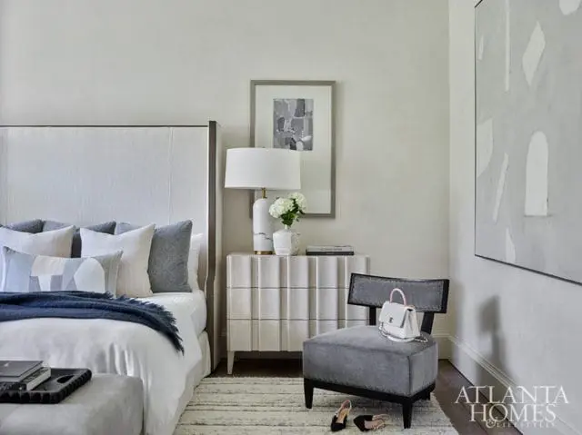 Bedroom with a white upholstered headboard, gray and white patterned pillows, a navy throw blanket, a fluted white nightstand with a white lamp and vase, and a gray accent chair with a white handbag.