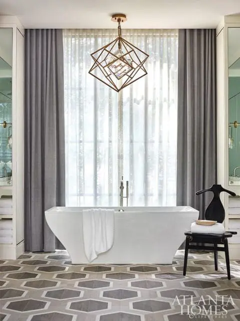 Modern bathroom with a freestanding white bathtub, geometric patterned floor, grey curtains, and a striking geometric chandelier.