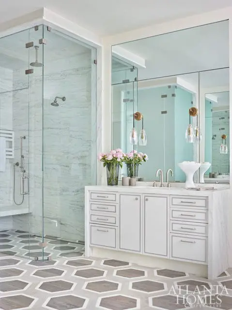 White bathroom vanity with marble countertop, double sinks, and mirrored cabinets. Features a glass-enclosed shower with marble tile walls and a hexagonal floor pattern.