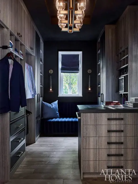 Walk-in closet with grey wood cabinetry, dark walls, blue velvet bench, and a modern chandelier. A navy suit hangs on a rack.
