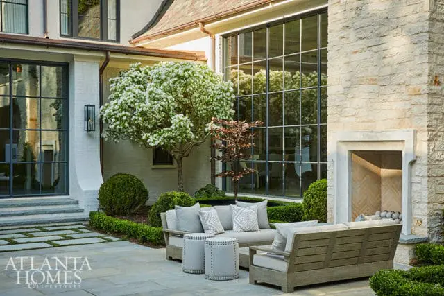 Stone patio with light gray outdoor sofa and chairs, two studded ottomans, and a blooming tree. Large grid windows reflect the outdoors.