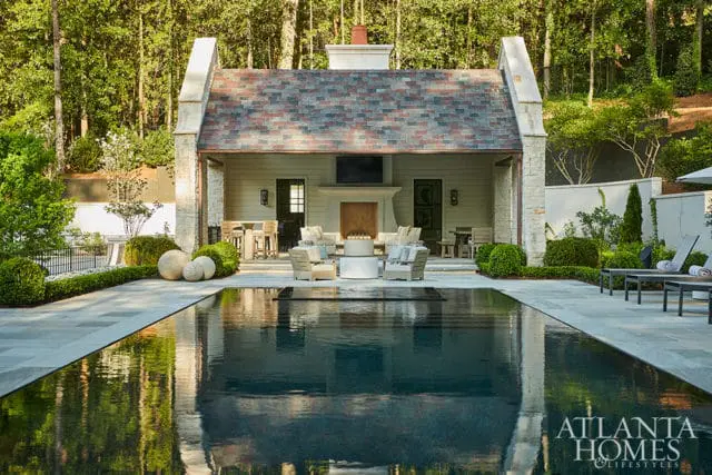 Modern outdoor living space with a dark-bottomed swimming pool, stone patio, and covered pavilion featuring a fireplace and seating area, surrounded by lush landscaping and trees.