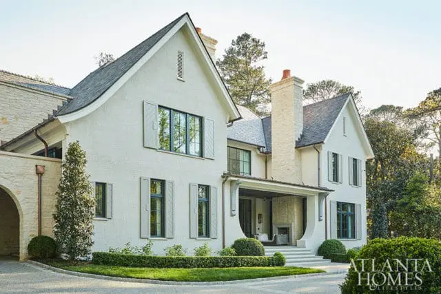 Modern home with white brick exterior, gray shutters, stone accents, and a slate roof. A covered porch with a fireplace is visible.