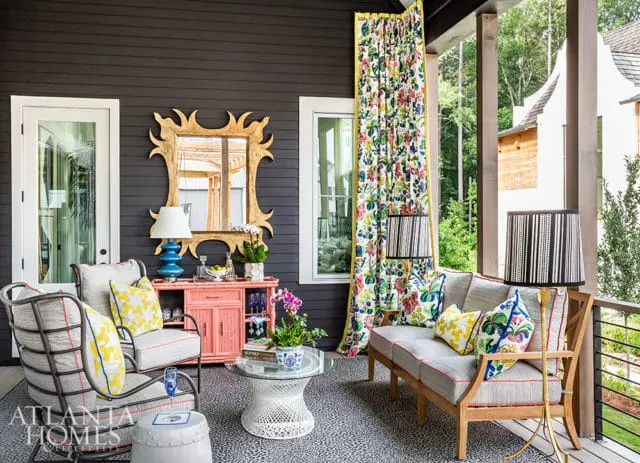Covered porch with dark gray siding, featuring two upholstered chairs and a sofa, a coral bar cabinet, a gold sunburst mirror, and floral curtains.