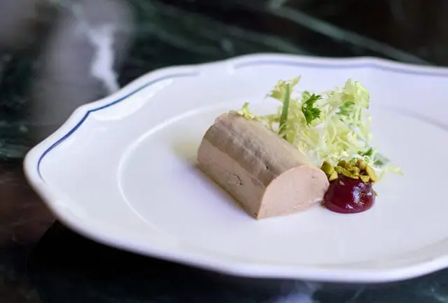 Terrine of pâté served with a small salad and a dollop of red berry compote topped with chopped pistachios on a white plate with a blue rim