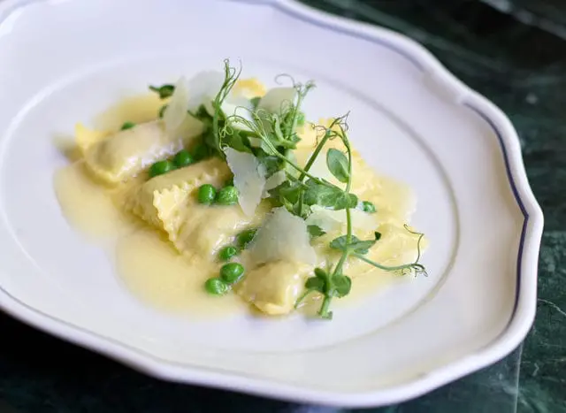 Ravioli with peas and shaved parmesan cheese, garnished with microgreens, served in a light butter sauce on a white plate with a blue rim.