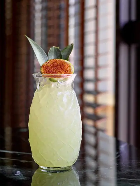 Pineapple-shaped glass filled with a pale yellow cocktail, garnished with a lime slice dusted with chili powder and pineapple leaves.