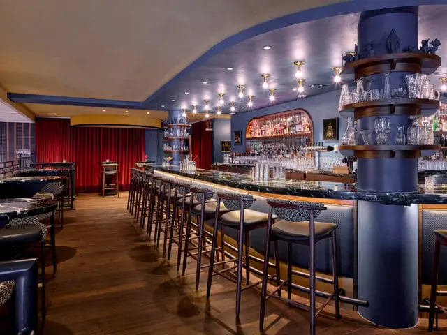 Bar with dark wood floors, blue walls, and a long marble-topped bar with upholstered stools. Red velvet curtains hang in the background.