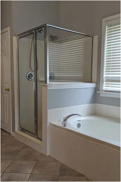Gray bathroom with a glass-enclosed shower, white tiled bathtub, and tiled floor. A white door is visible on the left.