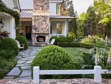 White house with stone chimney and outdoor fireplace, flagstone path, manicured hedges, and lush garden with foxglove and wooden obelisks.