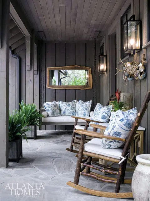 Covered porch with dark wood siding, a porch swing with blue and white floral cushions, two rustic rocking chairs with white cushions, and a live-edge mirror reflecting greenery.