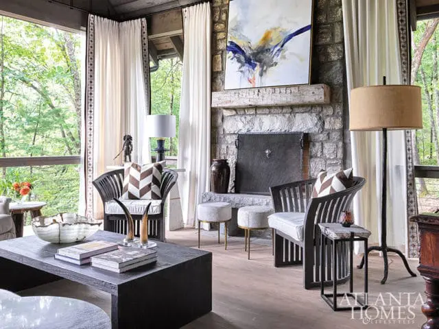 Living room with stone fireplace, dark wood chairs with patterned pillows, and large dark wood coffee table. Large windows overlook a wooded area.