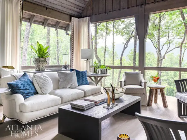 Living room with white sofa, blue and white patterned pillows, dark wood coffee table, and large windows overlooking a forest.