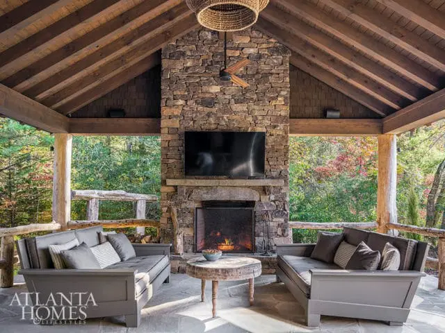 Covered outdoor living space with stone fireplace, television, two gray sofas, rustic wood accents, and a view of trees.