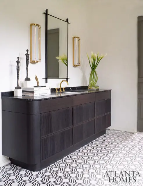 Dark wood vanity with fluted drawers, black marble countertop, and brass faucet, paired with geometric black and white floor tiles and gold sconces.
