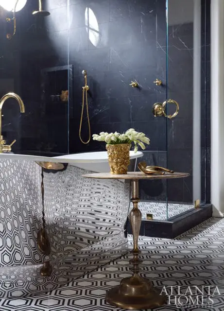 Master bathroom with black marble walls, freestanding tub with patterned skirt, gold fixtures, glass shower enclosure, and geometric floor tiles.