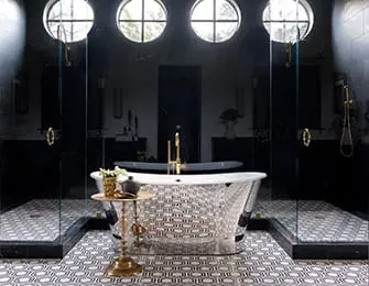 Modern bathroom with black marble walls, a freestanding clawfoot tub with gold fixtures, and glass-enclosed showers. The floor features a geometric tile pattern.