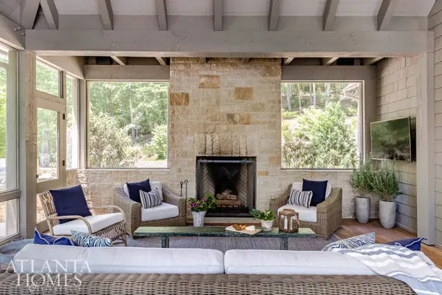 Screened porch with stone fireplace, wicker seating, and a sectional sofa. Large windows overlook a wooded area.